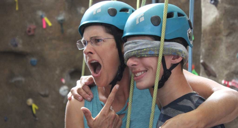 Two people who appear to have been rock climbing, based on their helmets and the rock wall in the background, embrace each other. One of them is blindfolded, and the other points, seeming impressed. 